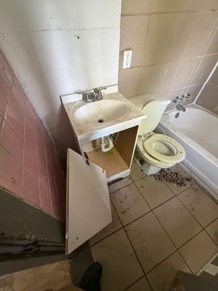 Bathroom as found: pink tile, a broken vanity with the door off its hinges and the floor filthy