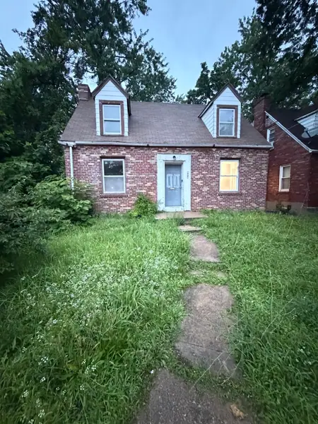 Brick cape before work: knee-high grass swallowing the front walk, overgrown shrubs against the foundation, weathered brick and a bare doorway