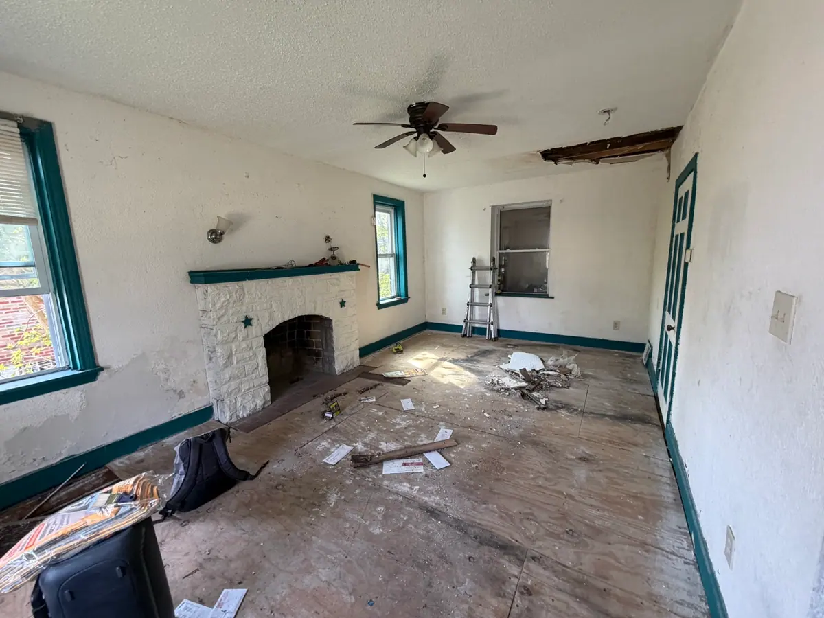 Living room stripped back: subfloor exposed, teal trim, a hole in the ceiling and debris across the room around a stone fireplace