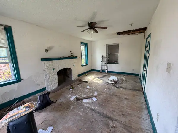 Living room stripped back: subfloor exposed, teal trim, a hole in the ceiling and debris across the room around a stone fireplace