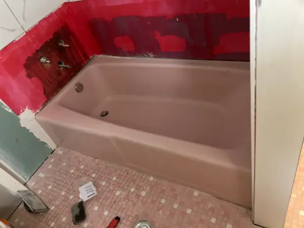 A dated pink cast-iron tub before refinishing, with the surround stripped back to red waterproofing and the valve rough-in exposed